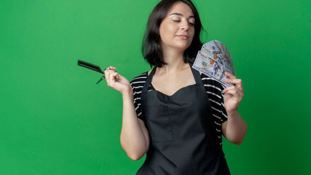 Hair stylist in a black apron holding hair-cutting tools and a fan of U.S. dollar bills.