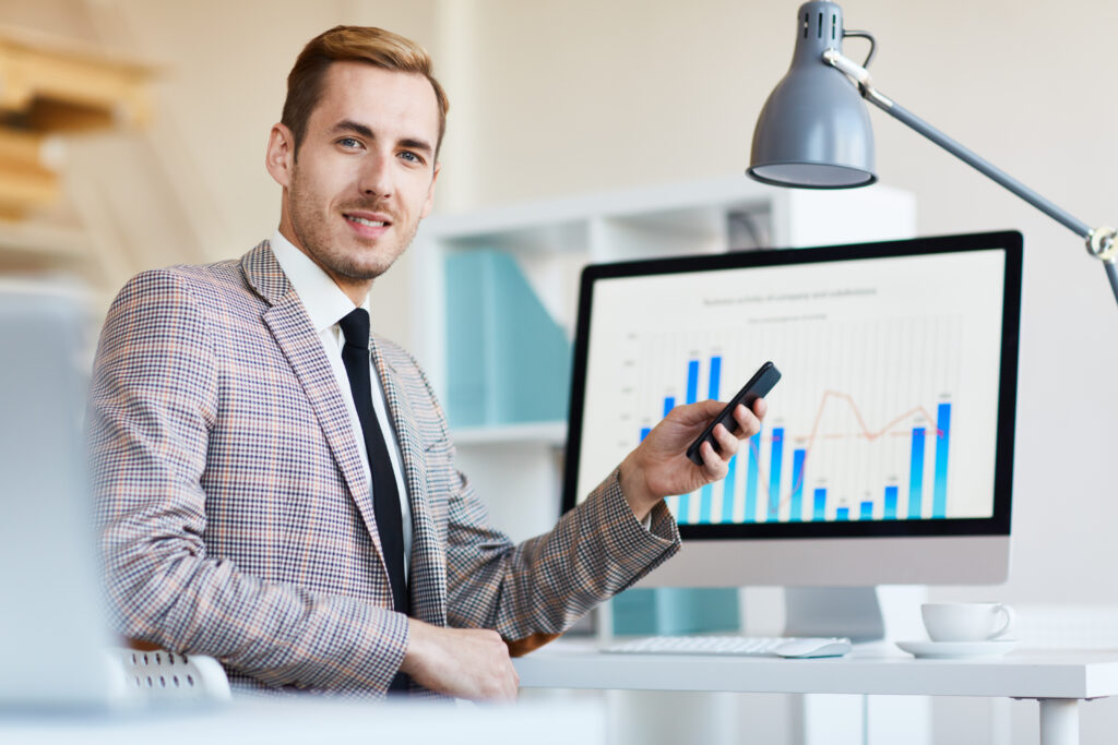 Businessman holding a smartphone in front of a desktop monitor showing analytics graphs.