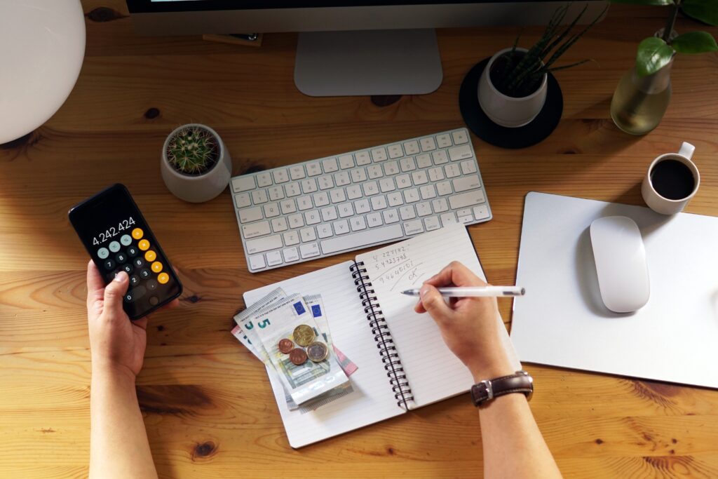 Person using a calculator app on a smartphone while writing budget notes beside Euro banknotes and coins.